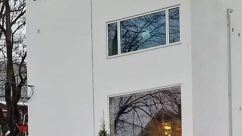 Hvit funkisbygning med store vinduer og åpen takterrasse, omgitt av vintervegetasjon.